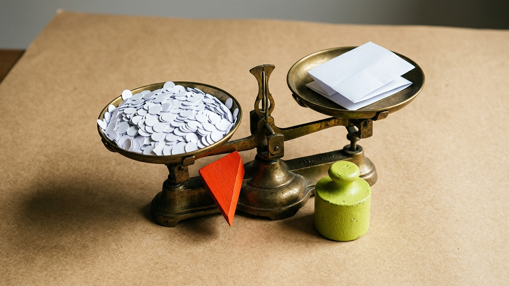 A brass scale weighs a heap of small paper discs against one folded payslip, tipped by a red wooden wedge, with a green counterweight beside it.